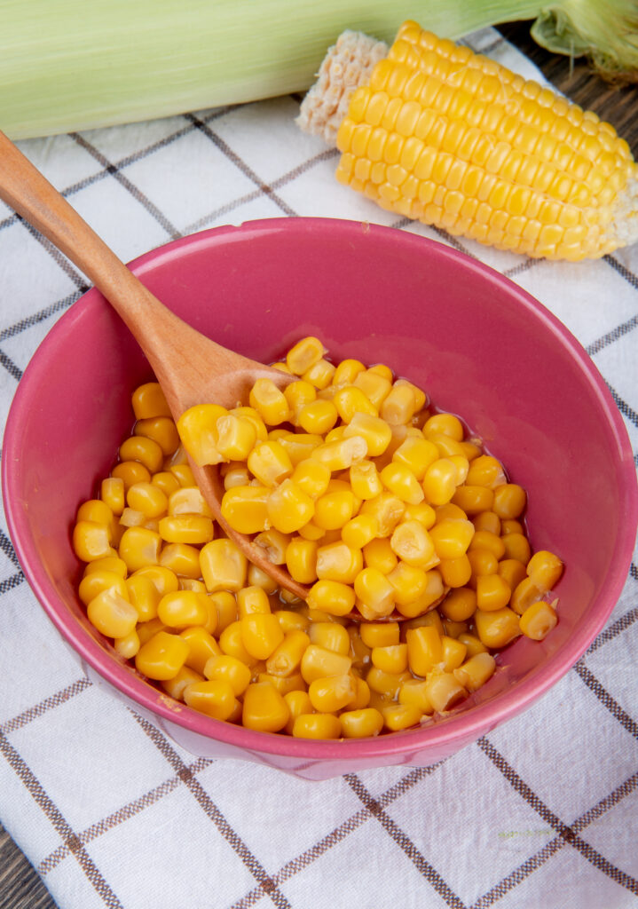 side view of bowl of yellow pea and wooden spoon with cut corn on plaid cloth background