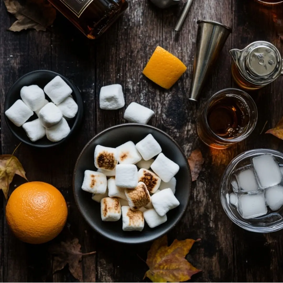 Flat lay of ingredients for toasted marshmallow Old Fashioned Angels Envy recipe including bourbon, marshmallows, bitters, and orange peel